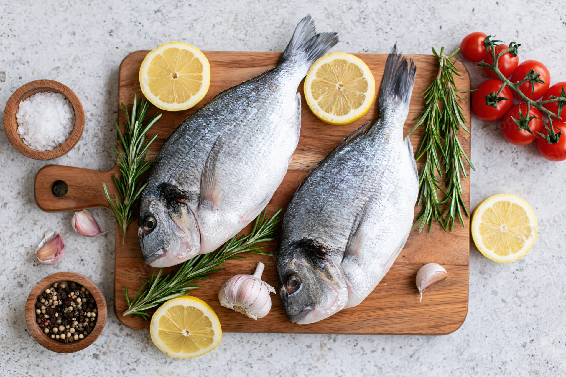 Two raw whole sea bream fish on a wooden cutting board with lemon, rosemary, garlic, tomatoes, salt and pepper arranged around them