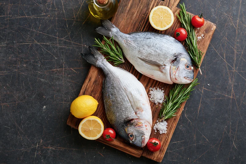 Two raw whole sea bream fish on a wooden cutting board with lemon, rosemary, tomatoes, salt, and olive oil on a dark countertop