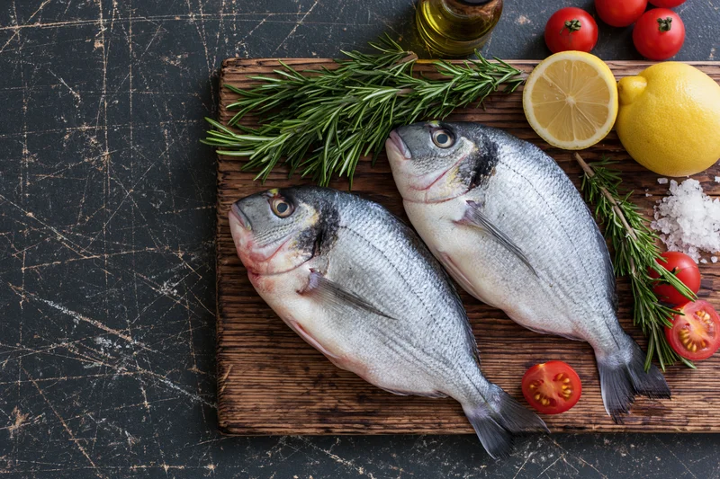 Two raw whole sea bream fish on a wooden cutting board with lemon, rosemary, tomatoes, and olive oil for meal preparation