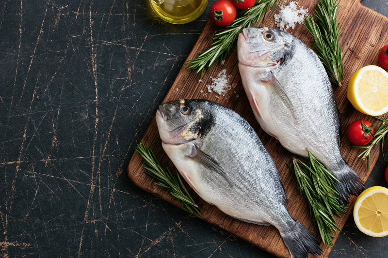 Two raw whole sea bream fish on a wooden cutting board with lemon, rosemary, tomatoes, and salt, set on a dark textured surface