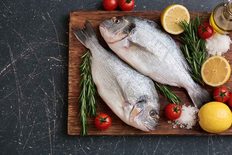Two raw whole sea bream fish on a wooden cutting board with lemon, rosemary, tomatoes, salt and olive oil for culinary preparation