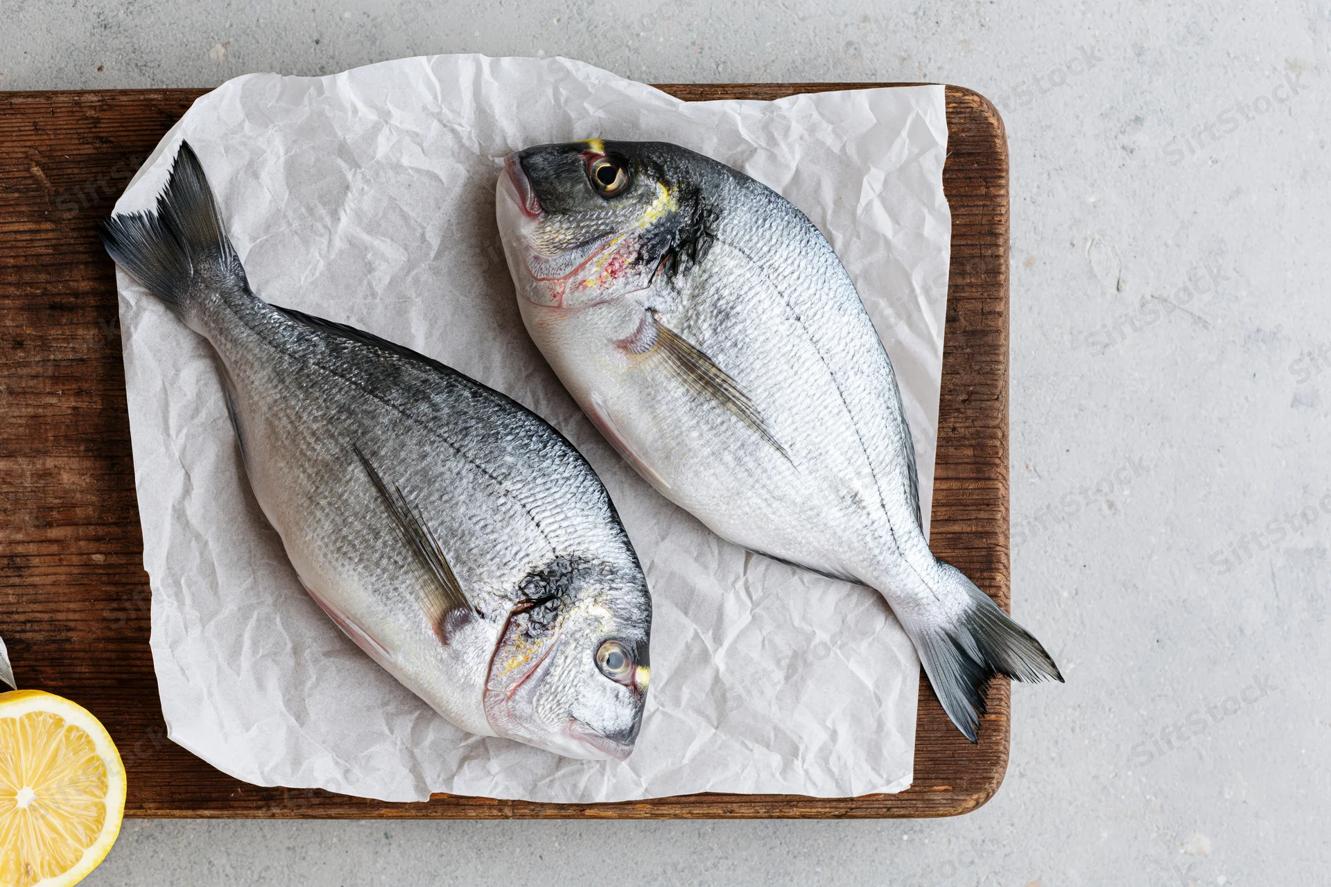 Two raw whole sea bream fish on crumpled parchment paper on a wooden cutting board with a lemon slice beside them