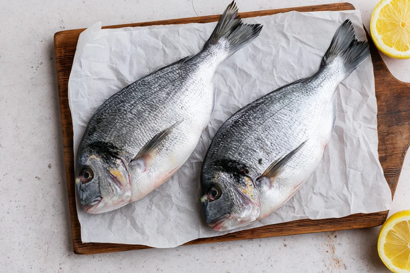 Two raw whole sea bream fish on parchment paper on a wooden cutting board with lemon slices nearby