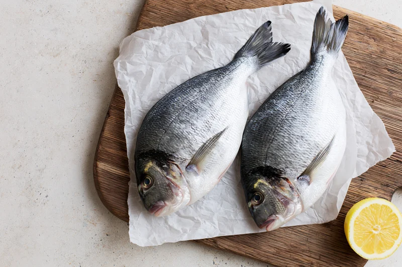 Two raw whole sea bream fish on parchment paper on a wooden cutting board with a lemon slice beside them