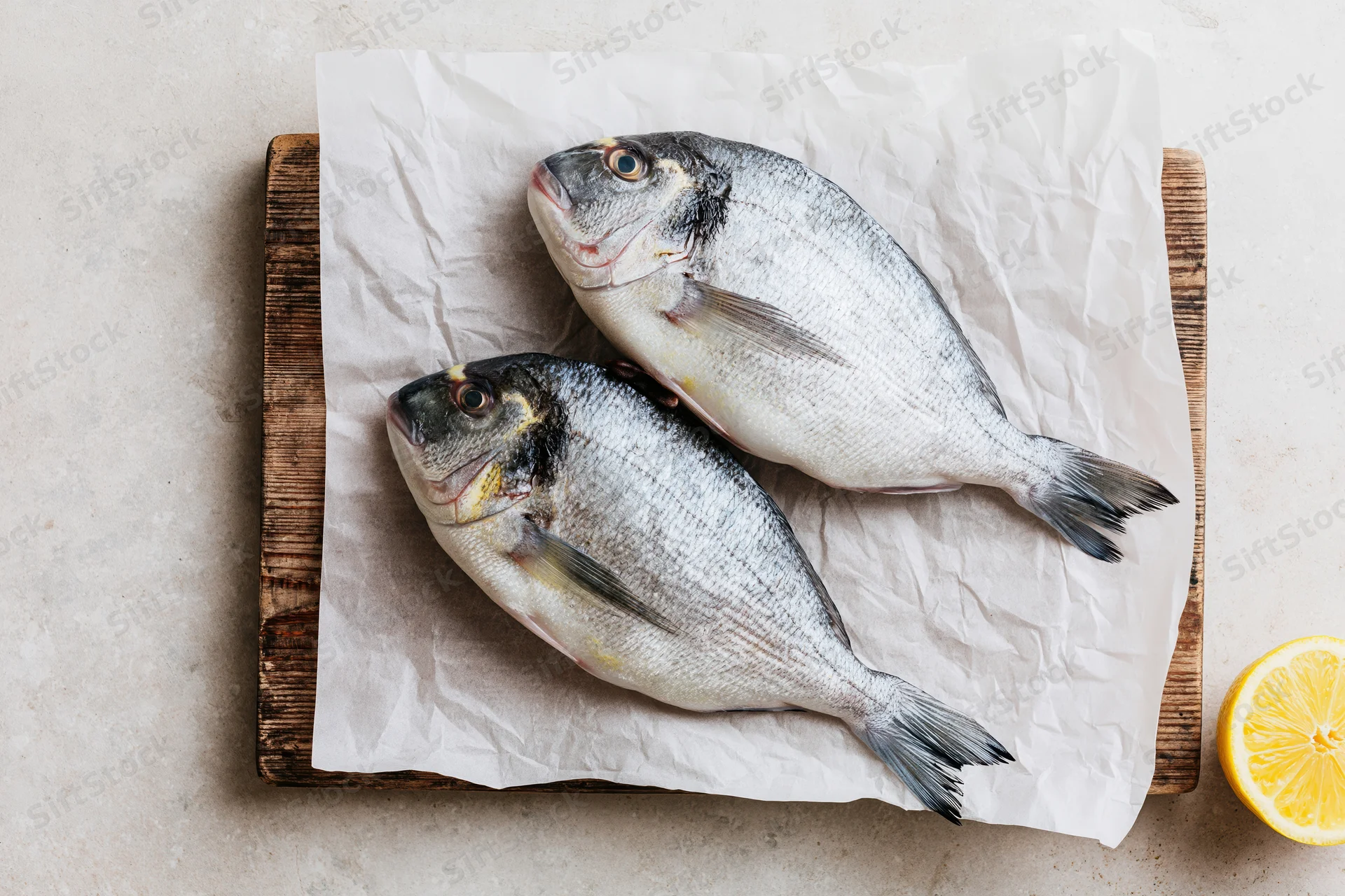 Two raw whole sea bream fish on crumpled parchment paper on a wooden cutting board with a lemon slice beside it