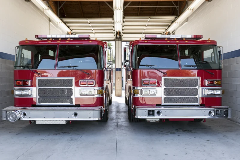 Two red fire trucks parked side by side inside an open fire station garage