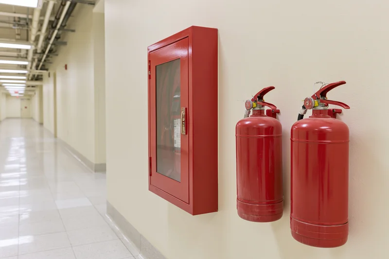 Two red fire extinguishers mounted on a beige wall in a long corridor with tiled floor and exposed ceiling pipes