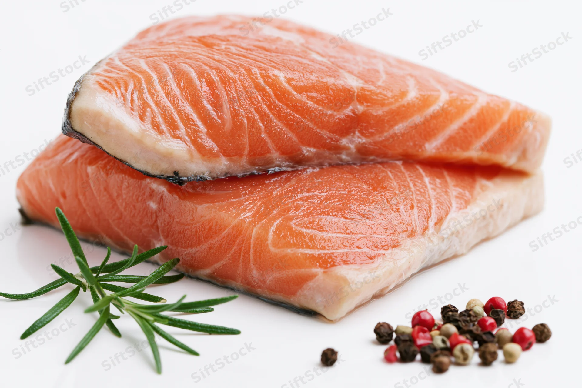 Two thick raw salmon fillets stacked on white background with rosemary and peppercorns