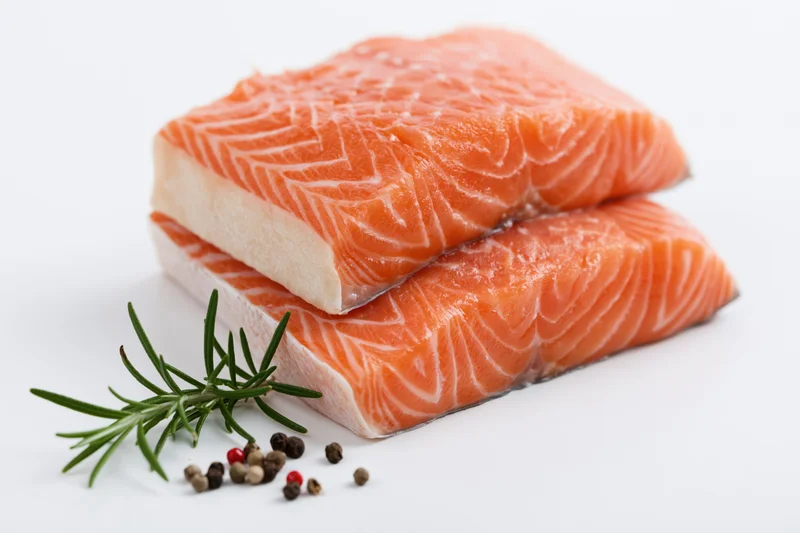 Two thick raw salmon fillets stacked with rosemary and peppercorns on white background