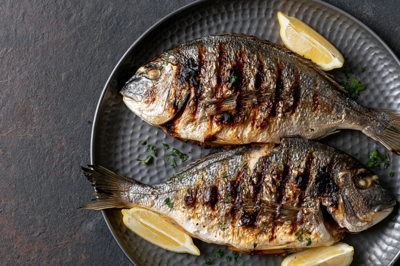 Two whole grilled sea bream fish on a dark hammered metal plate with lemon wedges and herbs
