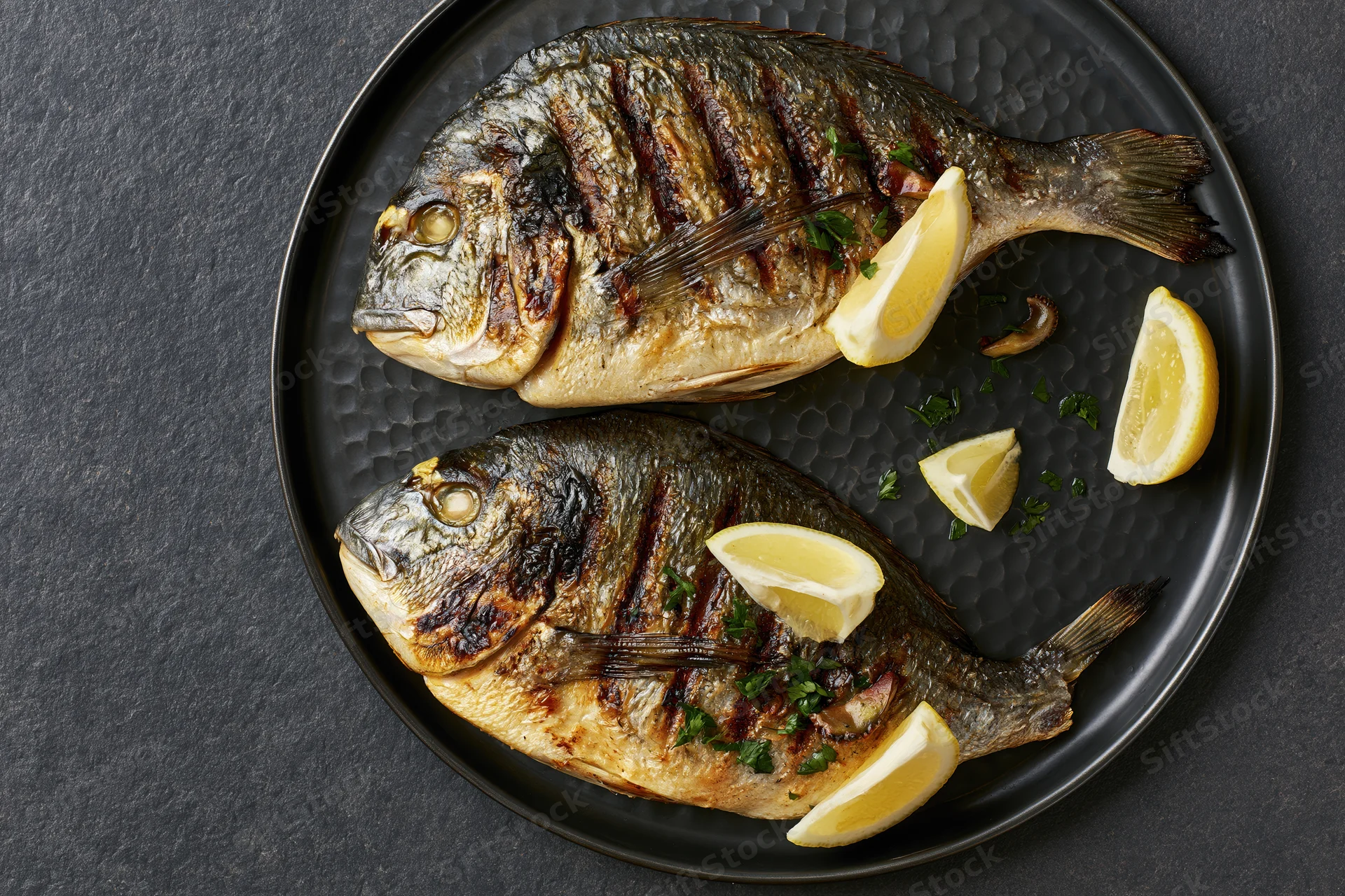 Two whole grilled sea bream fish on a dark hammered metal plate with lemon wedges and herbs