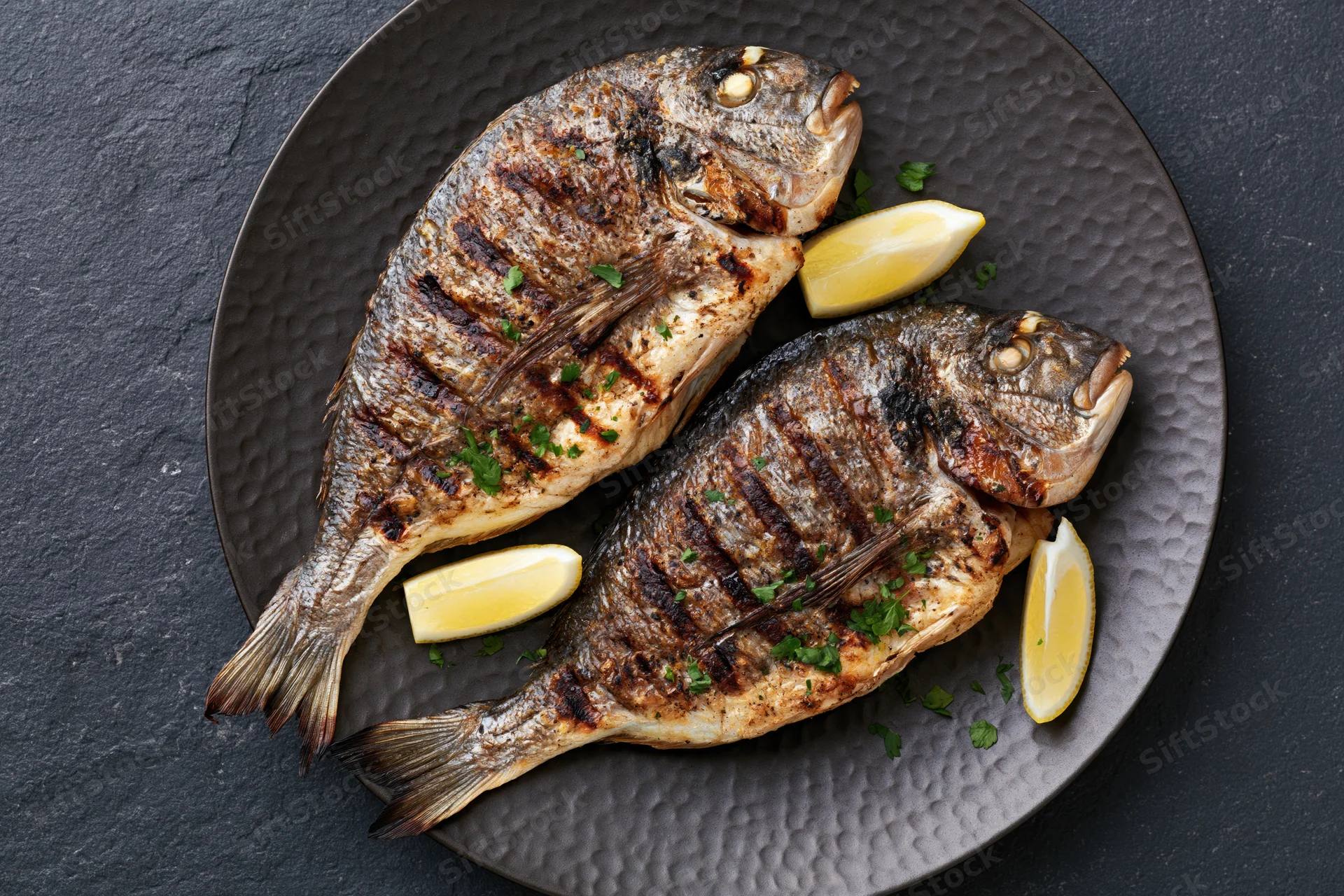 Two whole grilled sea bream fish on a dark hammered metal plate with lemon wedges and parsley garnish