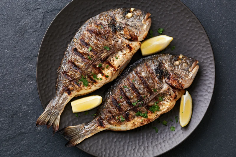 Two whole grilled sea bream fish on a dark hammered metal plate with lemon wedges and parsley garnish