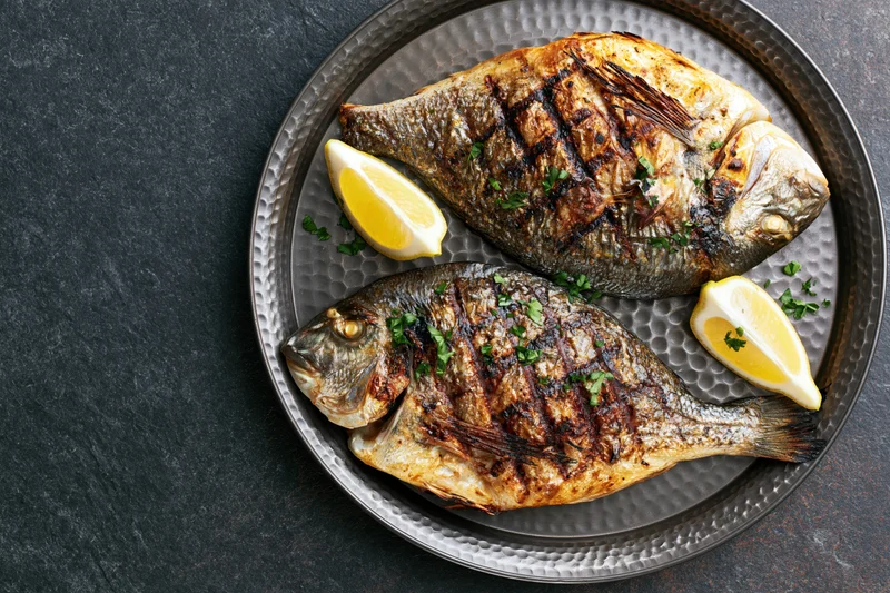 Two whole grilled sea bream fish on a dark hammered metal plate with lemon wedges and parsley garnish