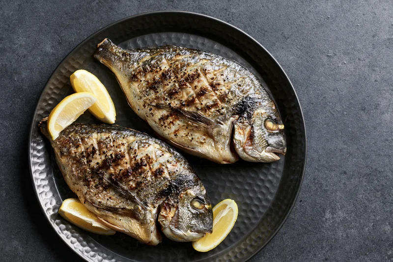 Two whole grilled sea bream fish on a dark hammered metal plate with lemon slices
