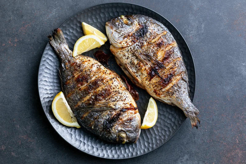 Two whole grilled sea bream fish with lemon slices on a dark hammered metal plate