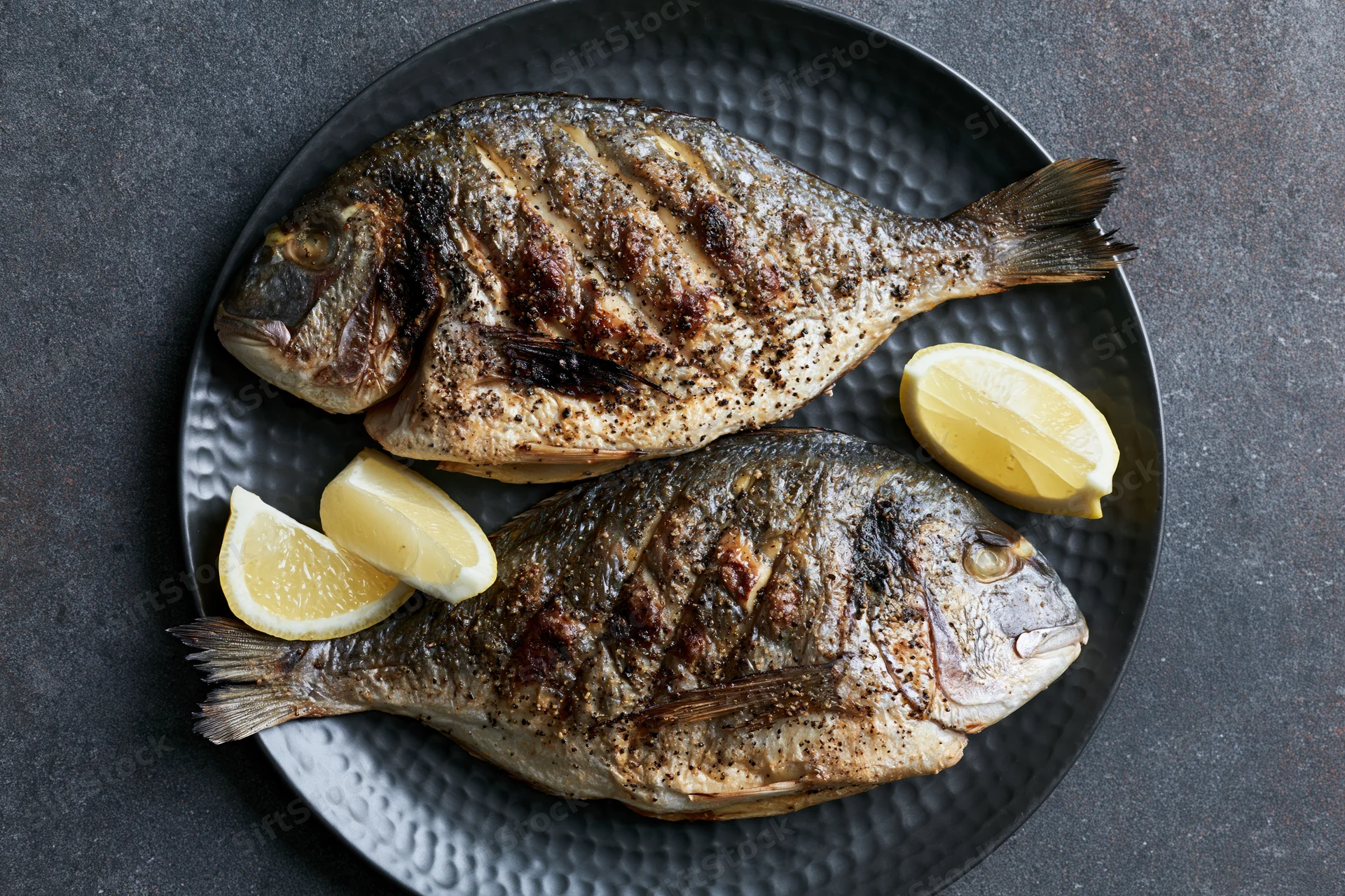 Two whole grilled sea bream fish on a dark hammered plate with lemon wedges, overhead view