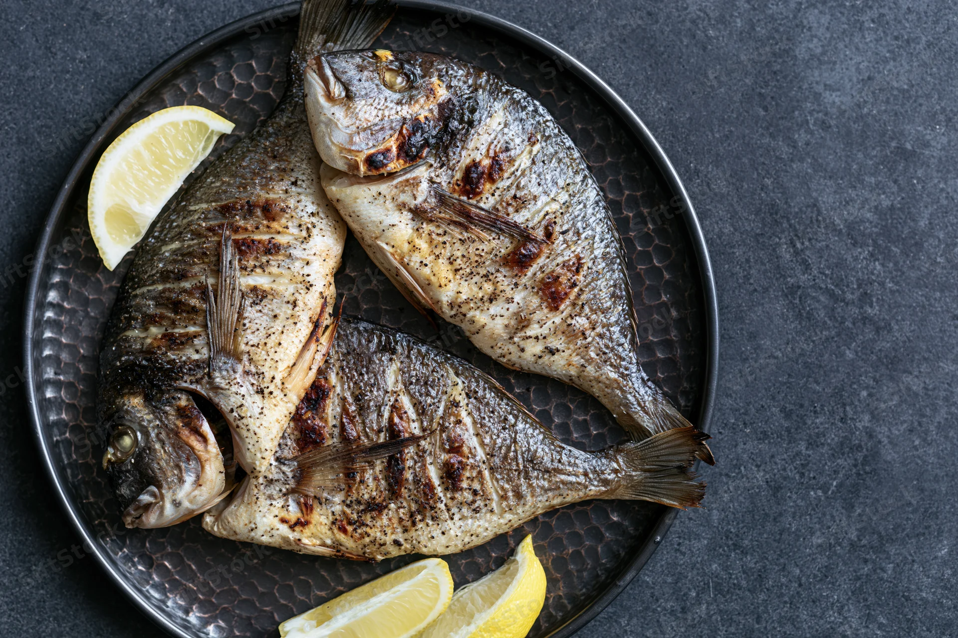Three whole grilled sea bream fish on a dark hammered metal plate with lemon slices