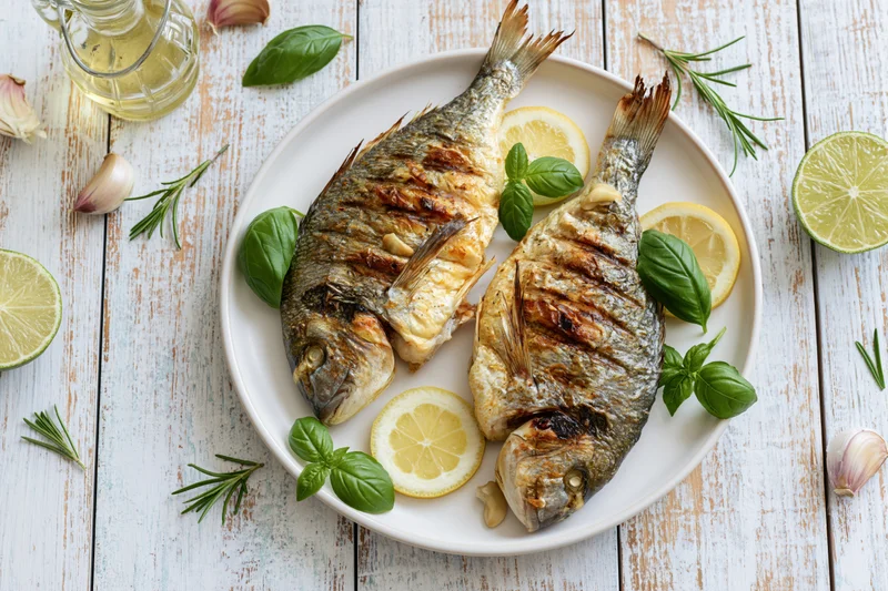 Two whole grilled sea bream fish on a white plate with lemon slices and herbs on a rustic wooden table
