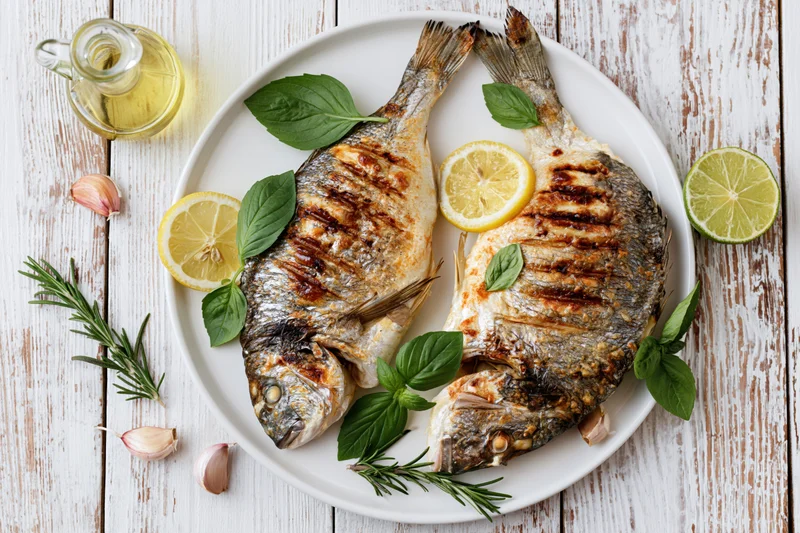 Two whole grilled sea bream fish on a white plate with lemon slices, basil, rosemary, garlic, and olive oil on a wooden table