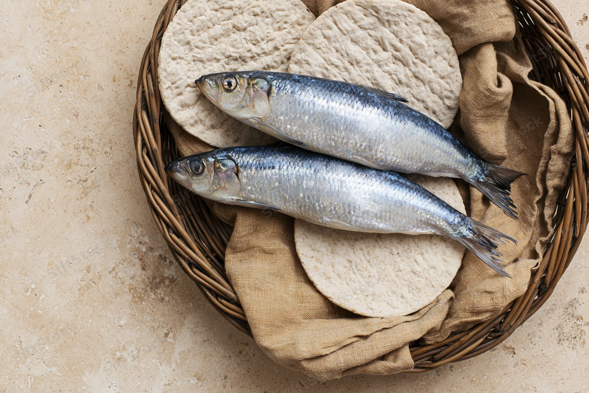 Two whole raw herring fish and round dimpled flatbreads arranged in a wicker basket with cloth lining