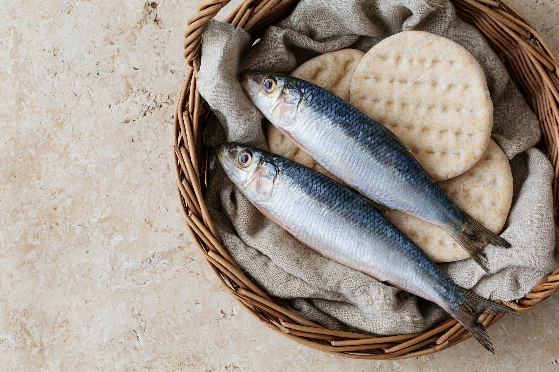 Two whole raw herring fish and round dimpled flatbreads in a wicker basket on a stone surface