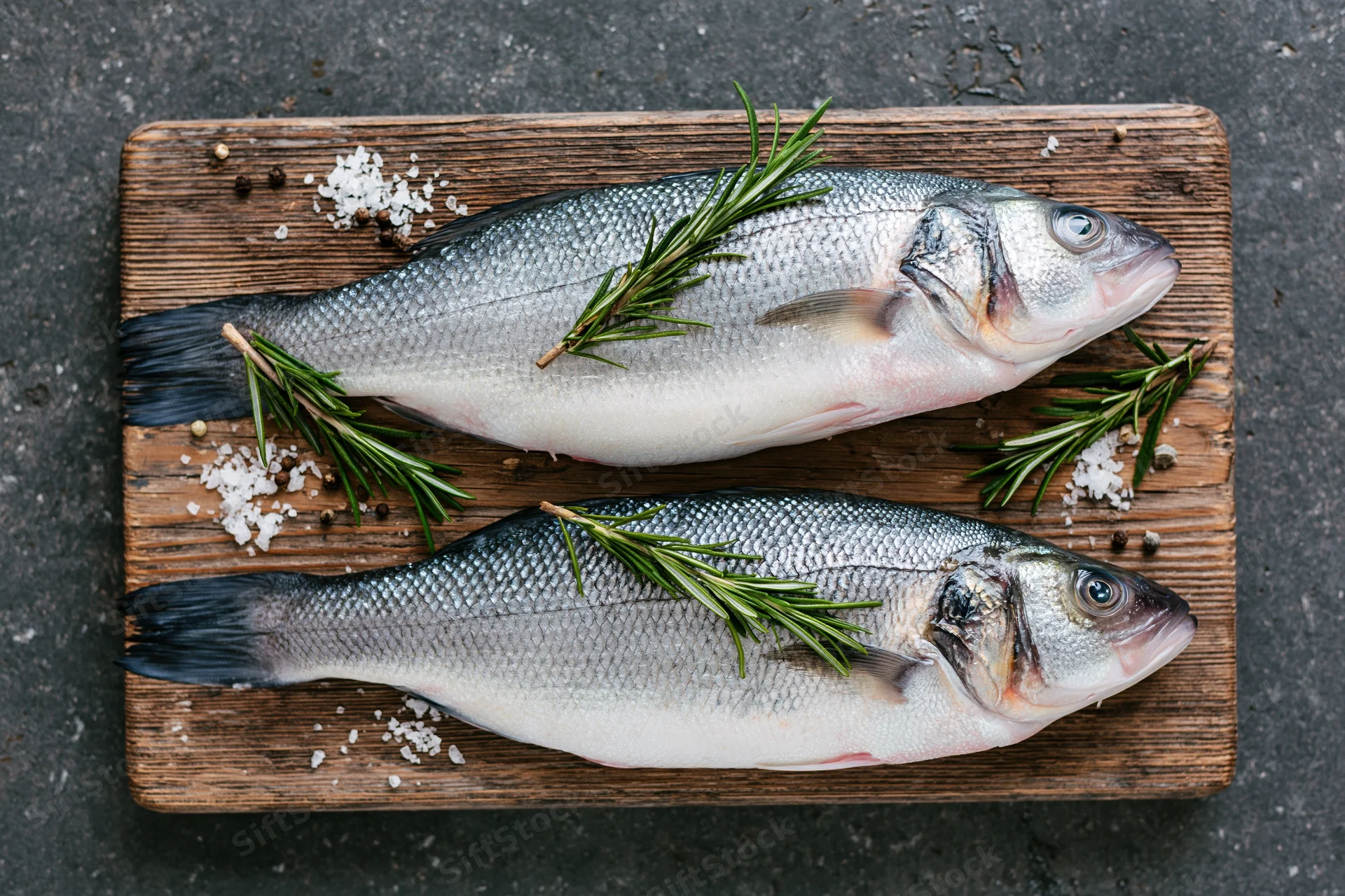 Two whole raw sea bass fish on a weathered wooden cutting board with rosemary, salt and pepper