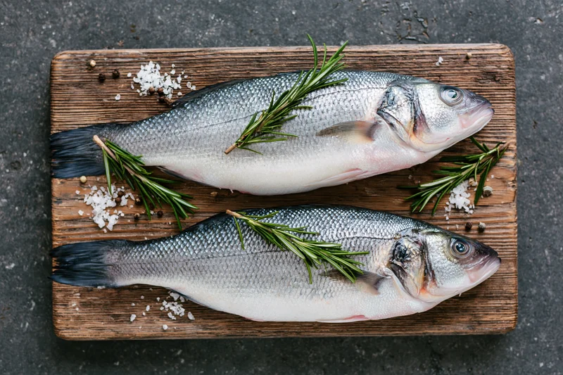 Two whole raw sea bass fish on a weathered wooden cutting board with rosemary, salt and pepper