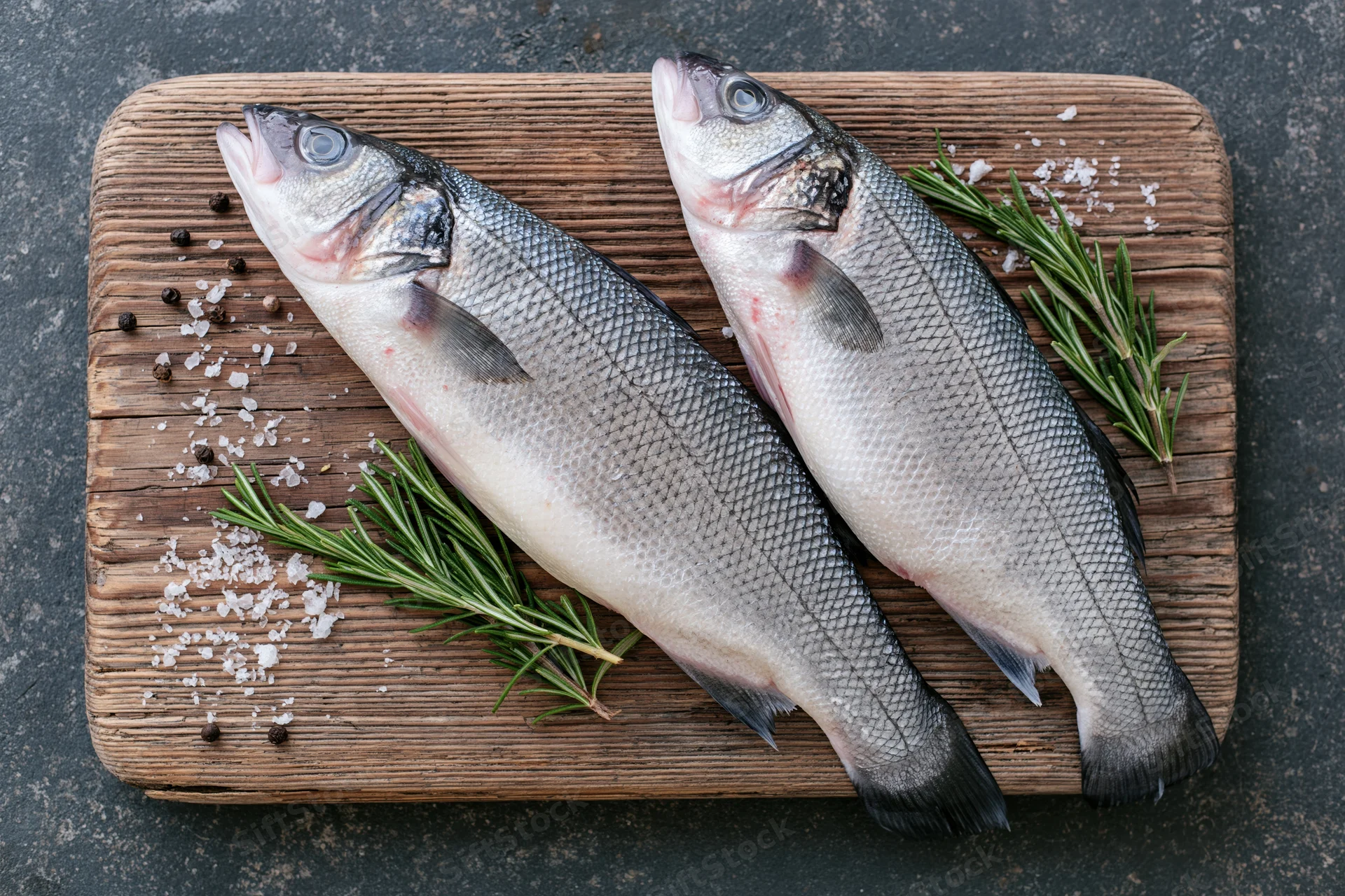 Two whole raw sea bass fish on a weathered wooden cutting board with rosemary, salt and pepper