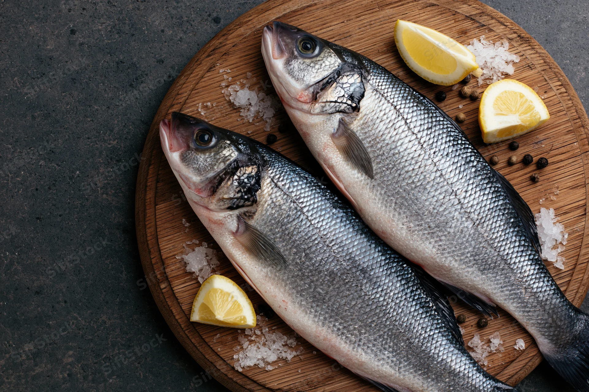 Two whole raw sea bass fish on a round wooden cutting board with lemon slices, salt, and pepper