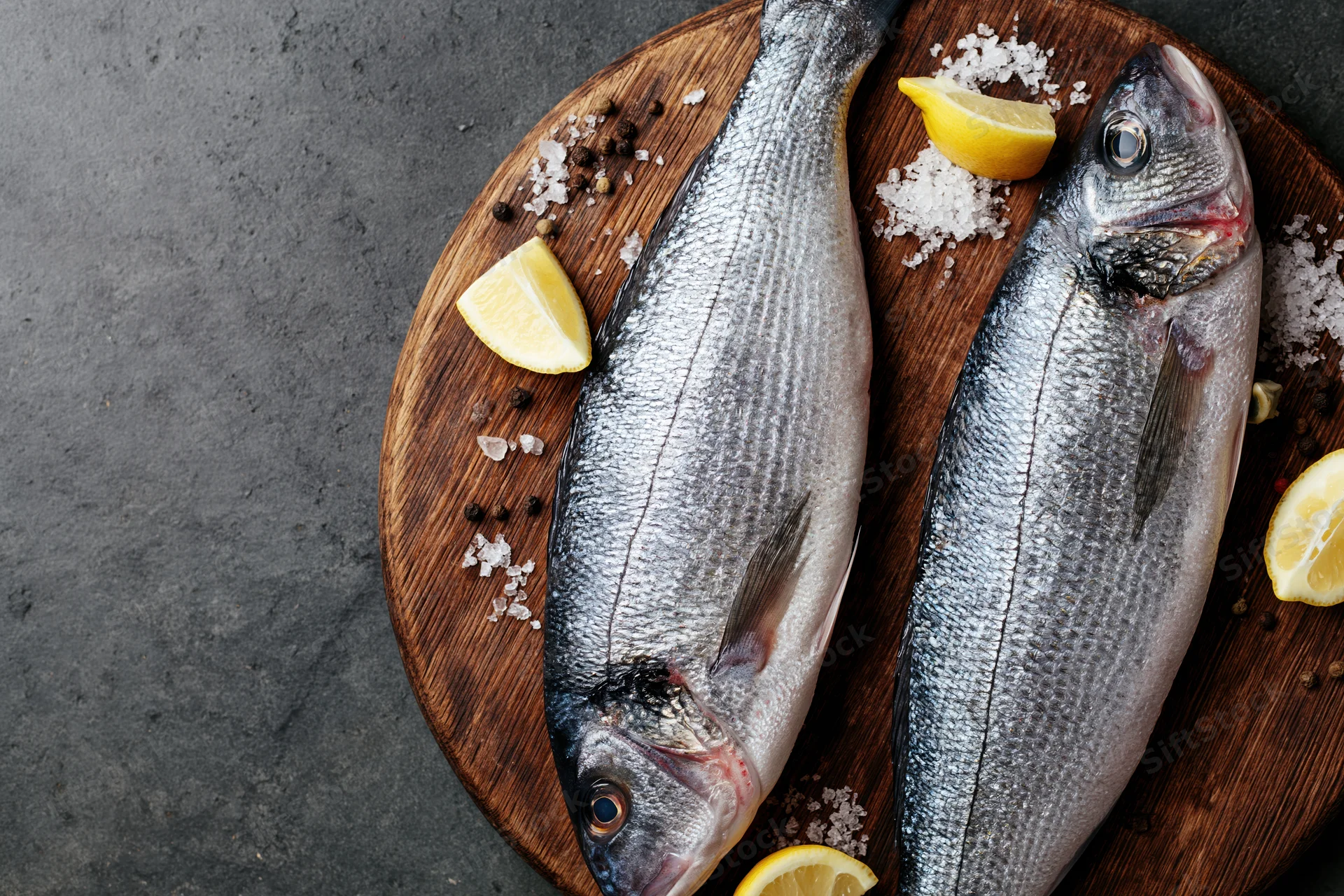 Two whole raw sea bass fish on a round wooden cutting board with lemon wedges and seasoning