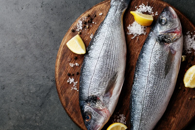 Two whole raw sea bass fish on a round wooden cutting board with lemon wedges and seasoning