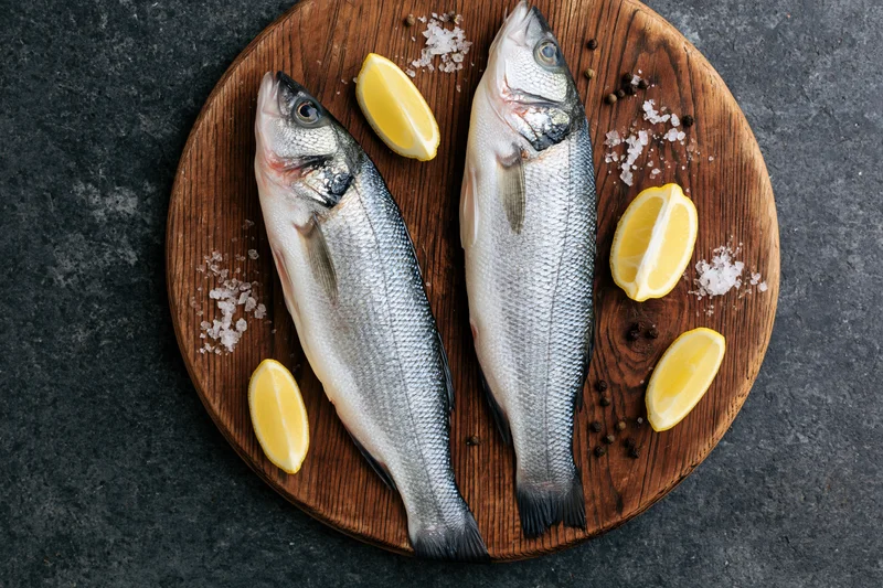 Two whole raw sea bass fish on a wooden board with lemon slices and seasoning, overhead view