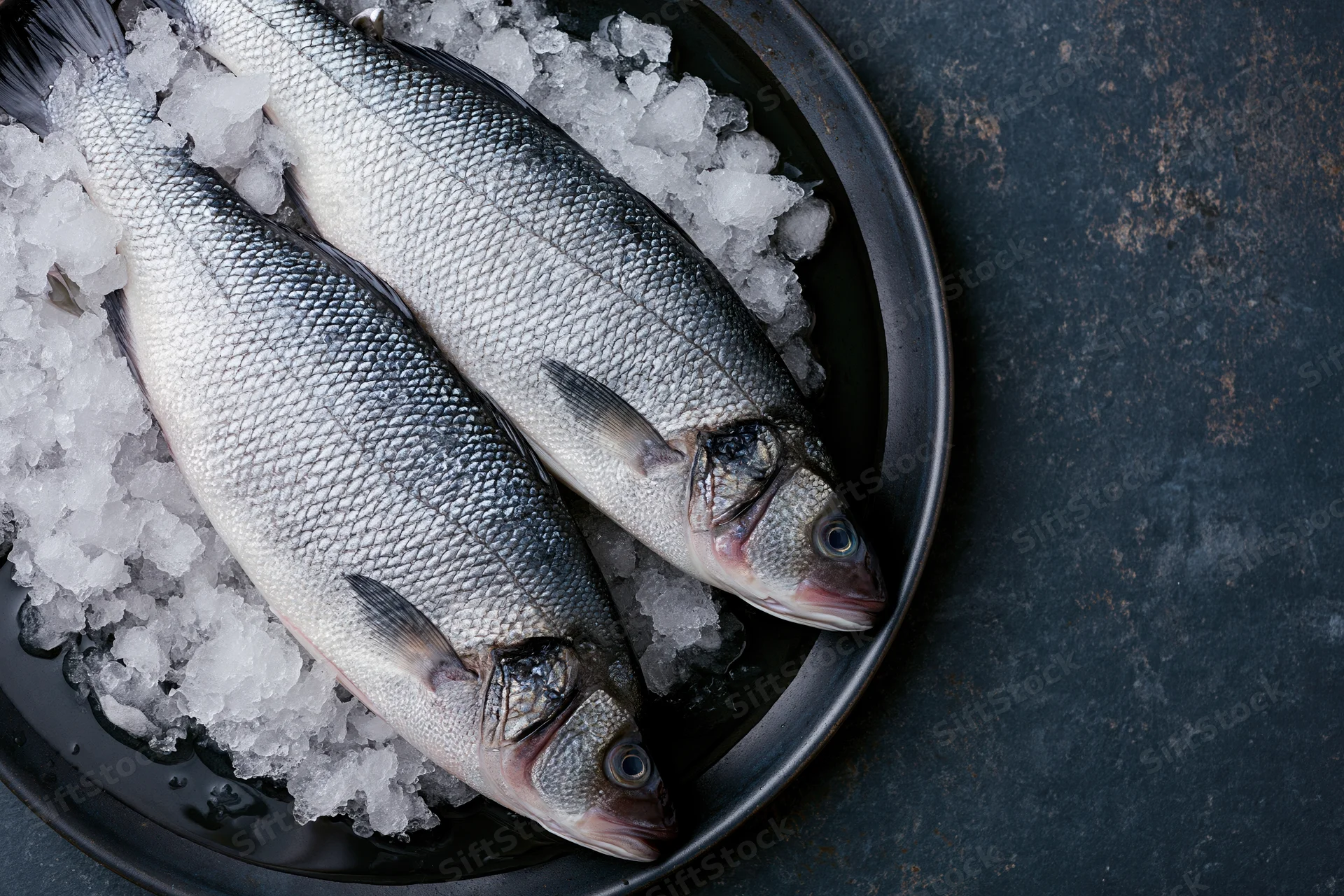 Two whole raw sea bass on crushed ice in a dark metal bowl
