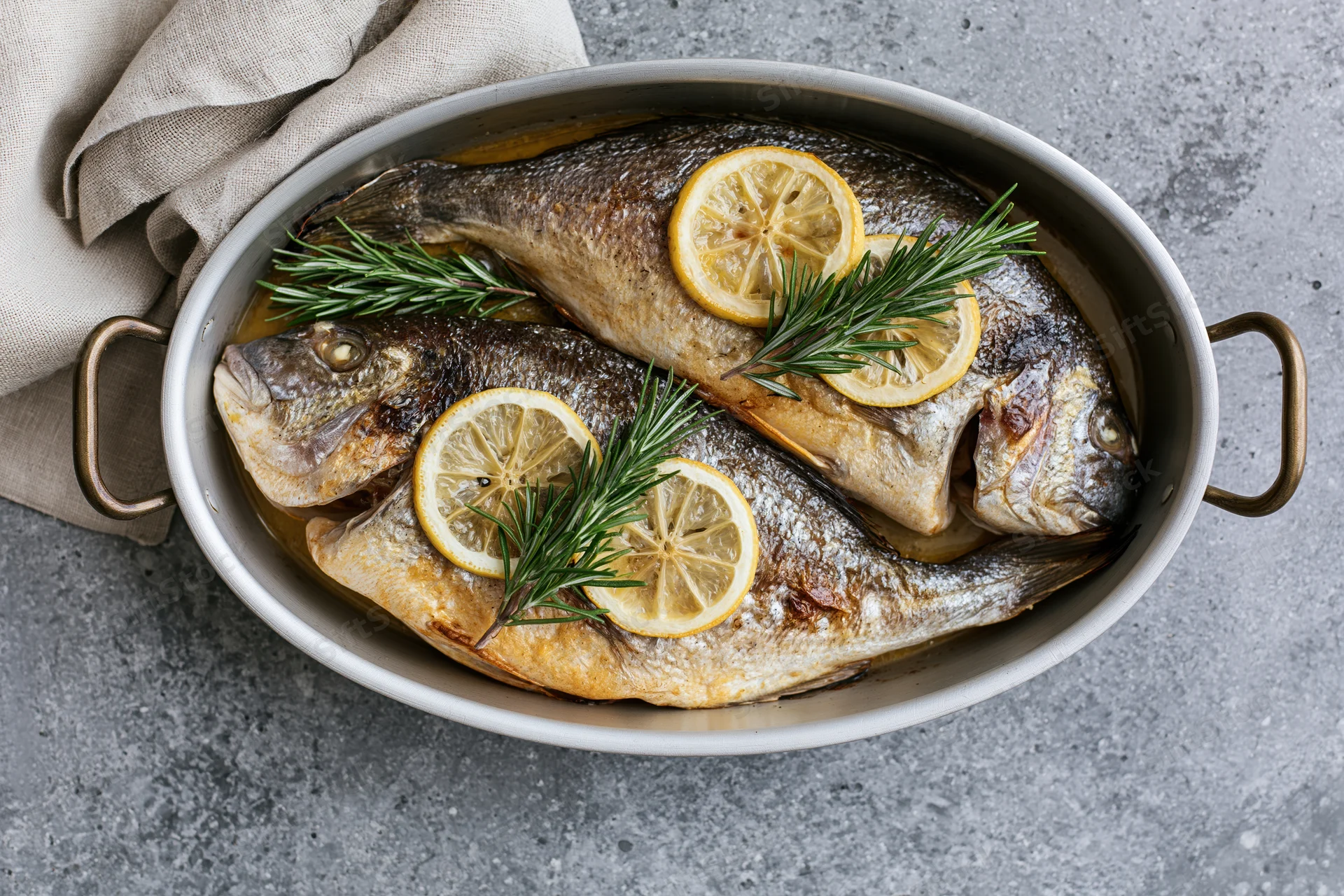 Two whole roasted sea bream fish in an oval metal baking dish garnished with lemon slices and rosemary sprigs on a gray countertop
