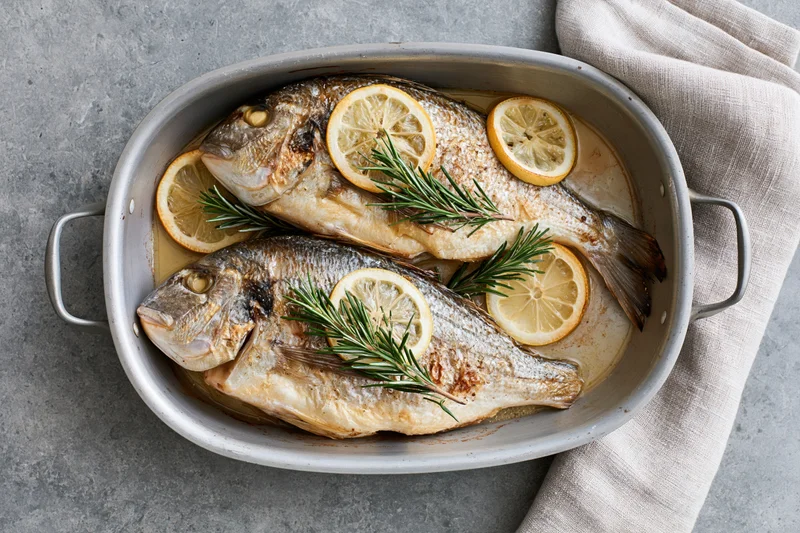 Two whole roasted sea bream fish in an oval metal baking dish with lemon slices and rosemary sprigs on a gray countertop