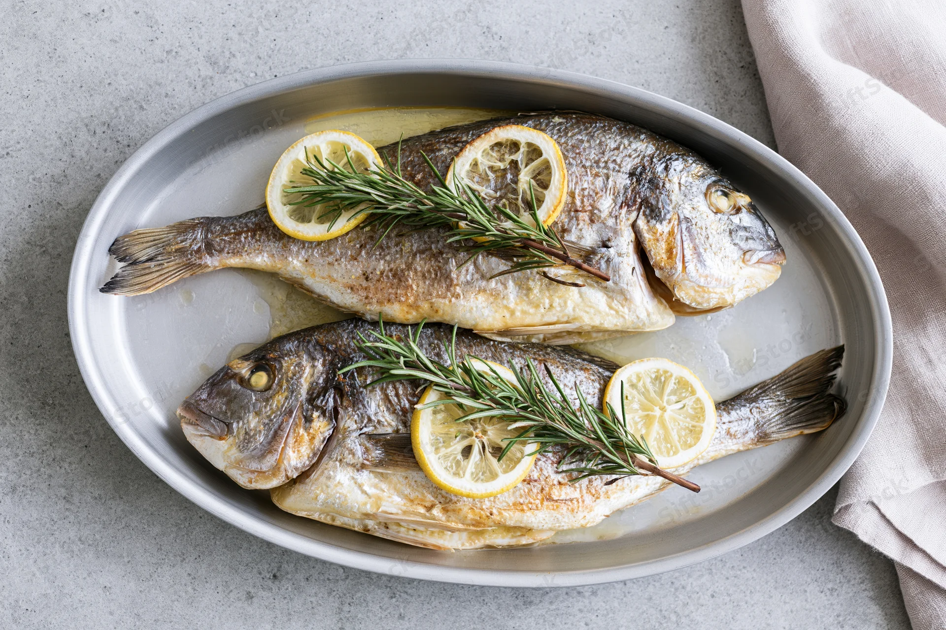 Two whole roasted sea bream fish in an oval metal baking dish garnished with lemon slices and rosemary sprigs on a gray countertop