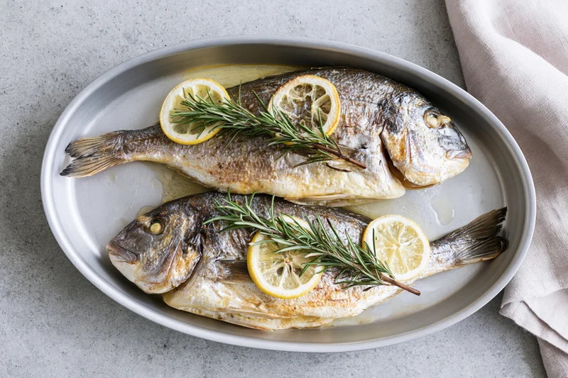 Two whole roasted sea bream fish in an oval metal baking dish garnished with lemon slices and rosemary sprigs on a gray countertop