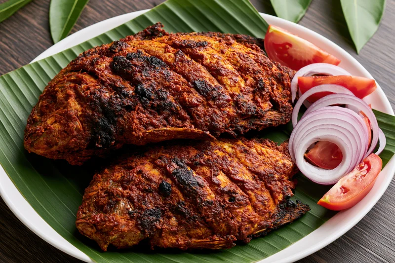 Two whole spiced fish with scored skin deep-fried and served on a banana leaf with tomato and onion garnish on a white plate