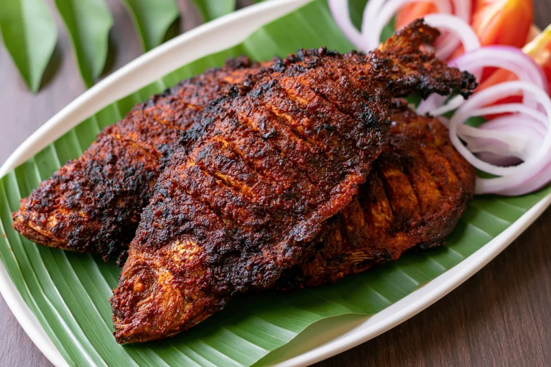 Two whole spiced fish with scored skin fried to crispy dark brown served on banana leaf in oval plate with sliced onions and tomatoes