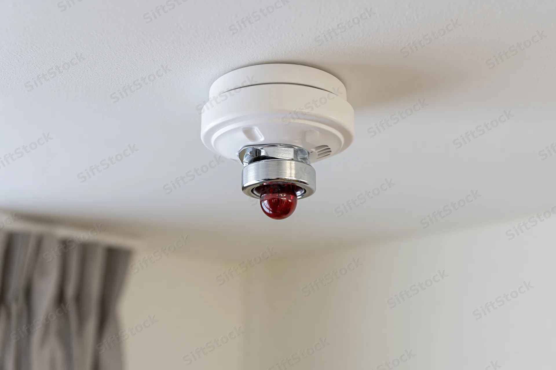 White smoke detector with red light and chrome sprinkler head mounted on ceiling in residential interior