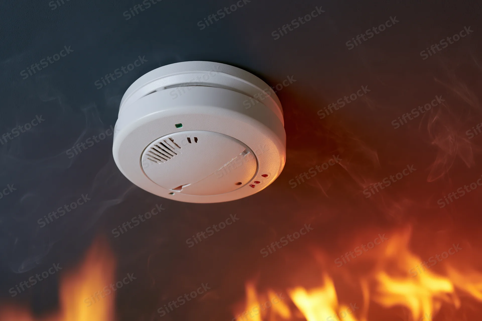 White circular smoke detector mounted on dark ceiling with flames and smoke below
