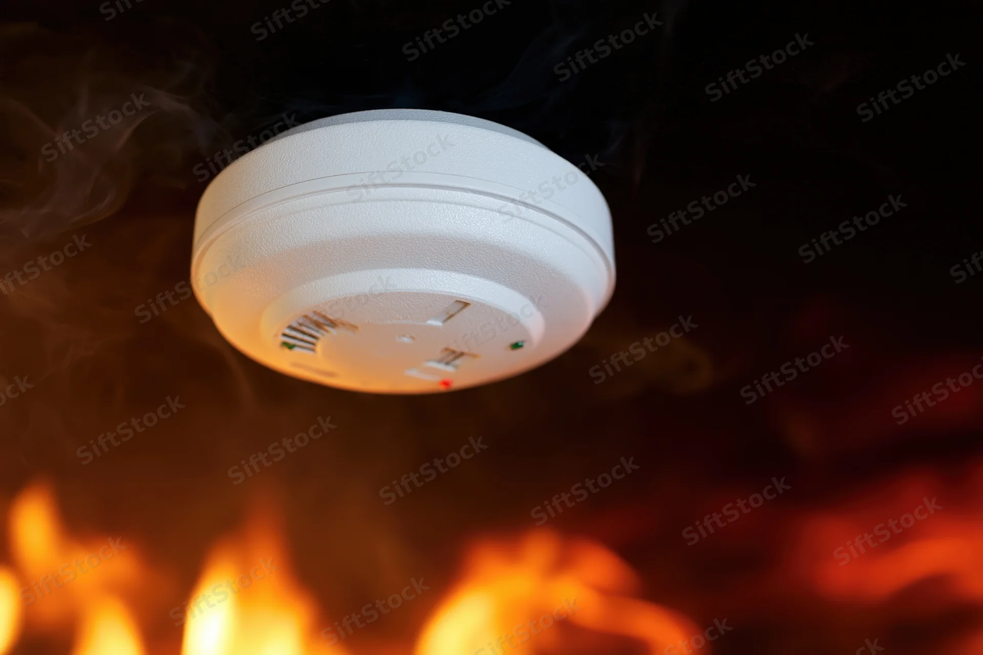 White circular smoke detector mounted on dark ceiling with flames and smoke below