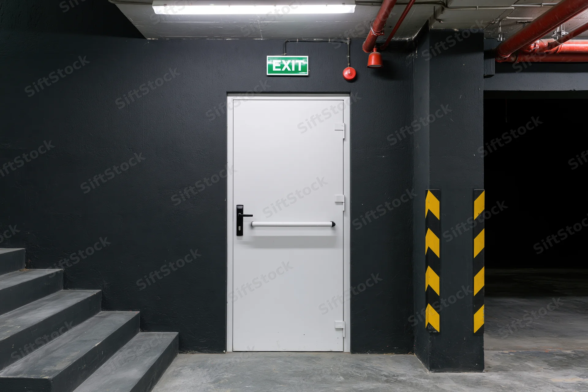 White metal fire exit door with push bar handle centered in a dark industrial hallway beside a staircase