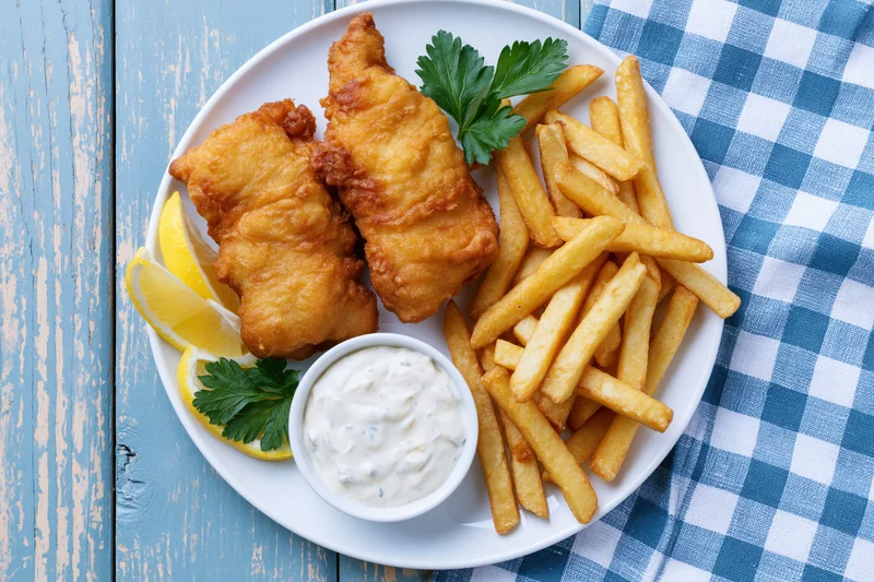 Battered fish and chips on a white plate with tartar sauce and lemon slices on a blue wooden table with checkered cloth