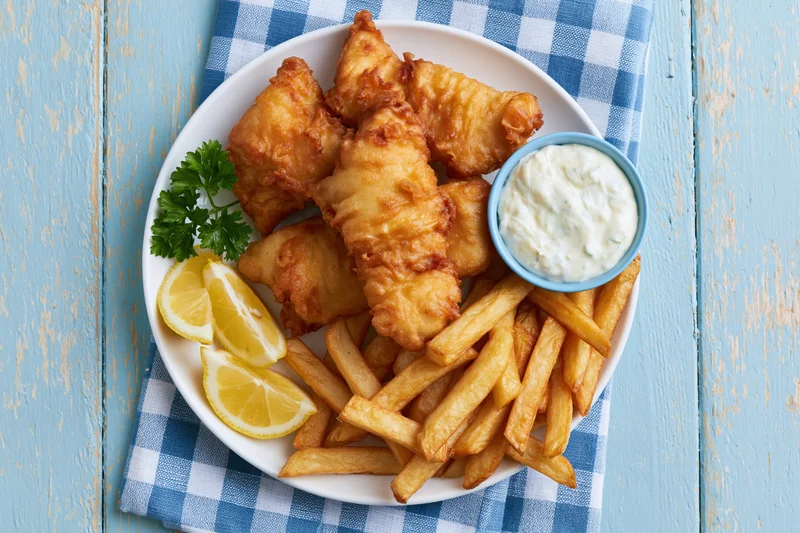 Battered fish and chips served on a white plate with lemon wedges and tartar sauce on a blue checkered napkin over a rustic wooden table