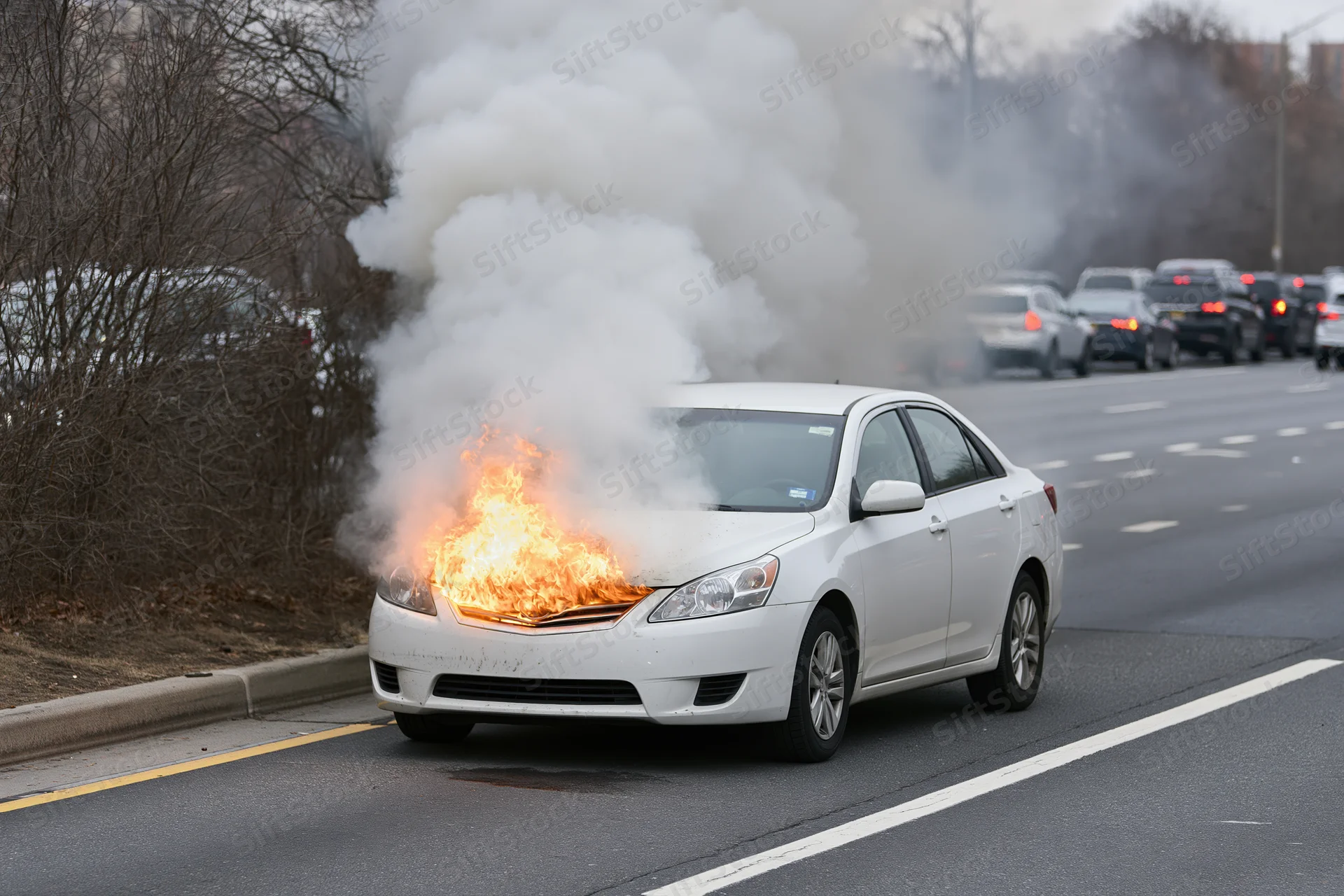 White sedan on fire with thick smoke on the shoulder of a multi-lane road