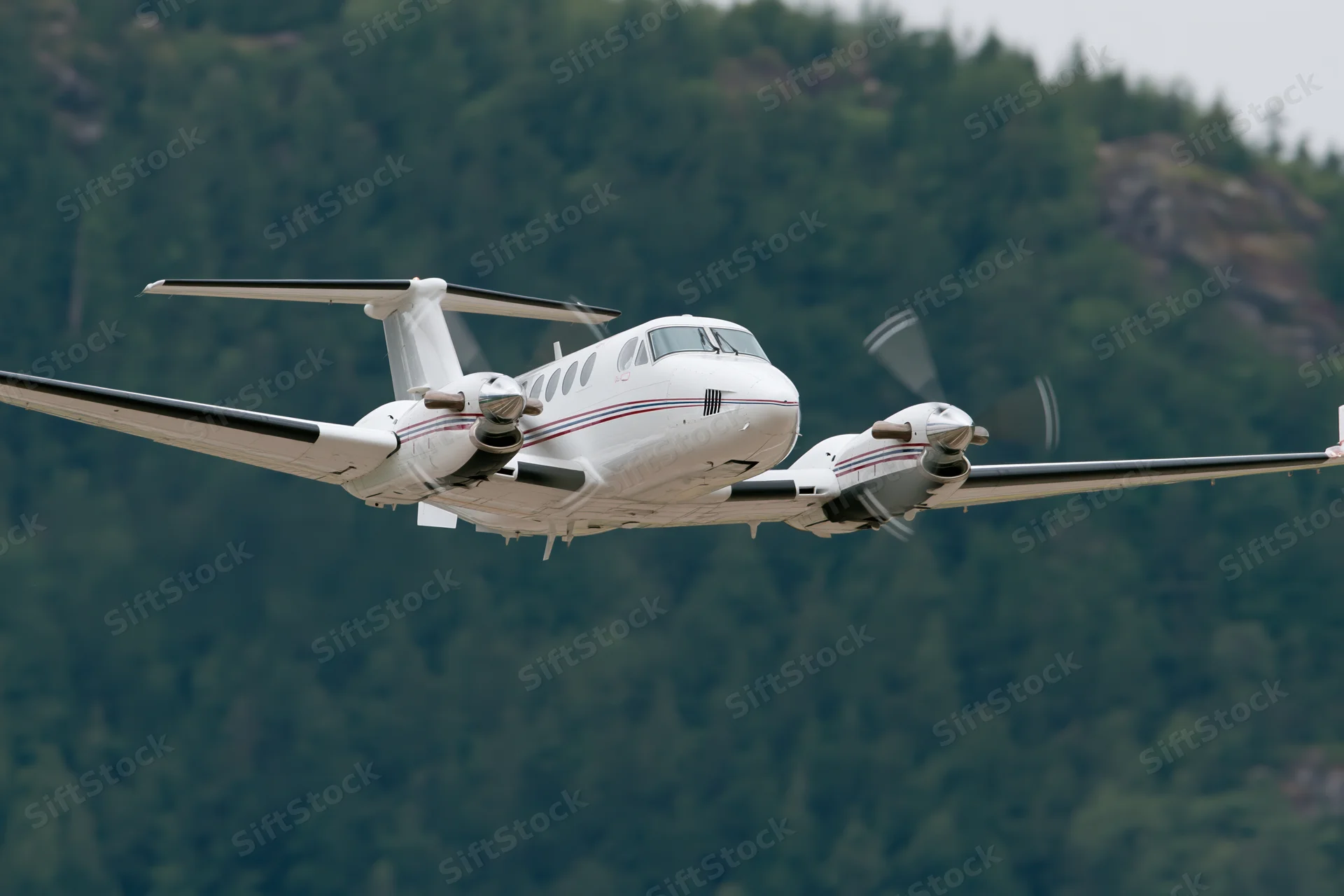 White twin-engine turboprop aircraft in low-altitude flight over forested mountains