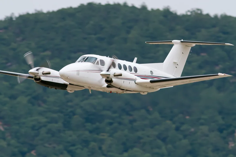 White twin-engine turboprop aircraft in low-altitude flight over forested mountains
