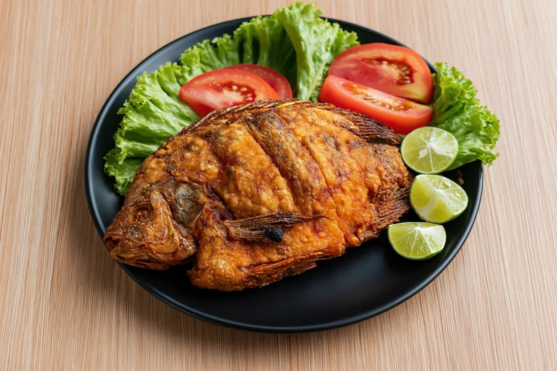 Whole fried tilapia with crispy golden-brown skin on a black plate with lettuce, tomato, and lime slices on a wooden table